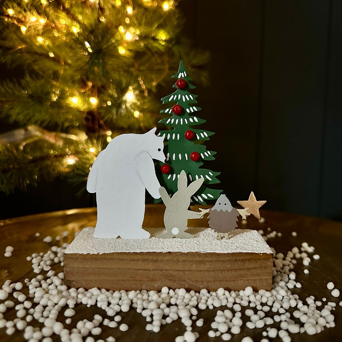 A handmade metal Christmas cutout scene featuring polar bear figure, rabbit and hedgehog, a decorated green Christmas tree, and a small star, displayed on a wooden base with white pebbles, with a blurred Christmas tree in the background