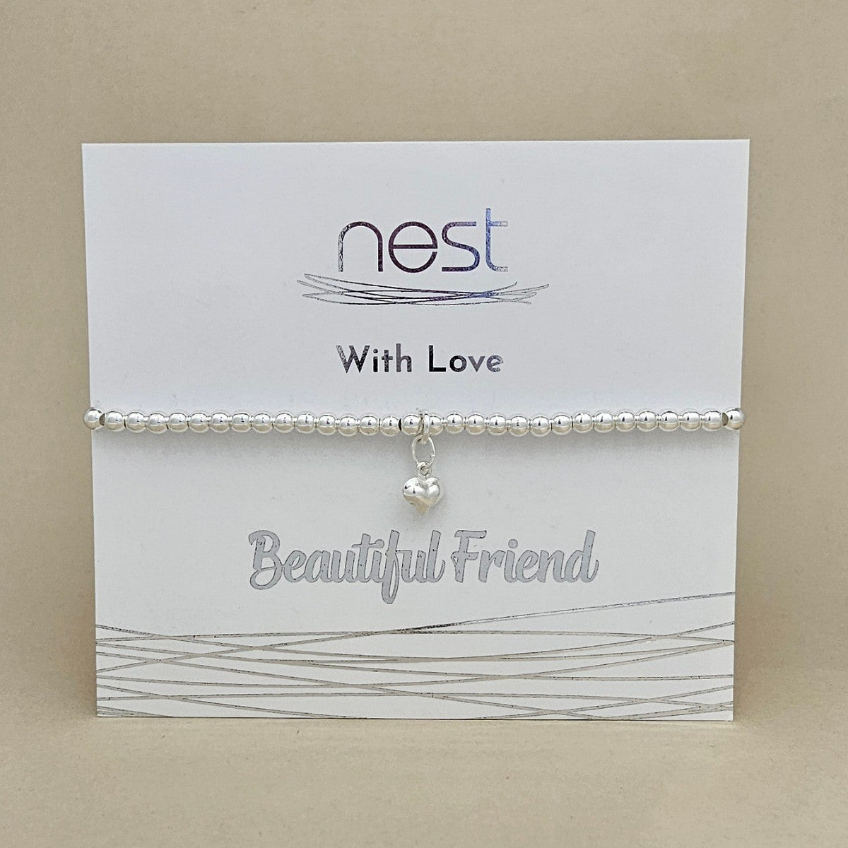 A silver beaded bracelet with a small dangling heart charm, displayed on a white branded card that reads 'nest With Love Beautiful Friend' against a beige background.