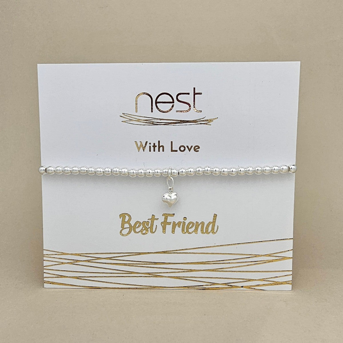 A silver beaded bracelet with a small dangling heart charm, displayed on a white card that reads 'nest With Love Best Friend' with gold decorative lines, set against a beige background.