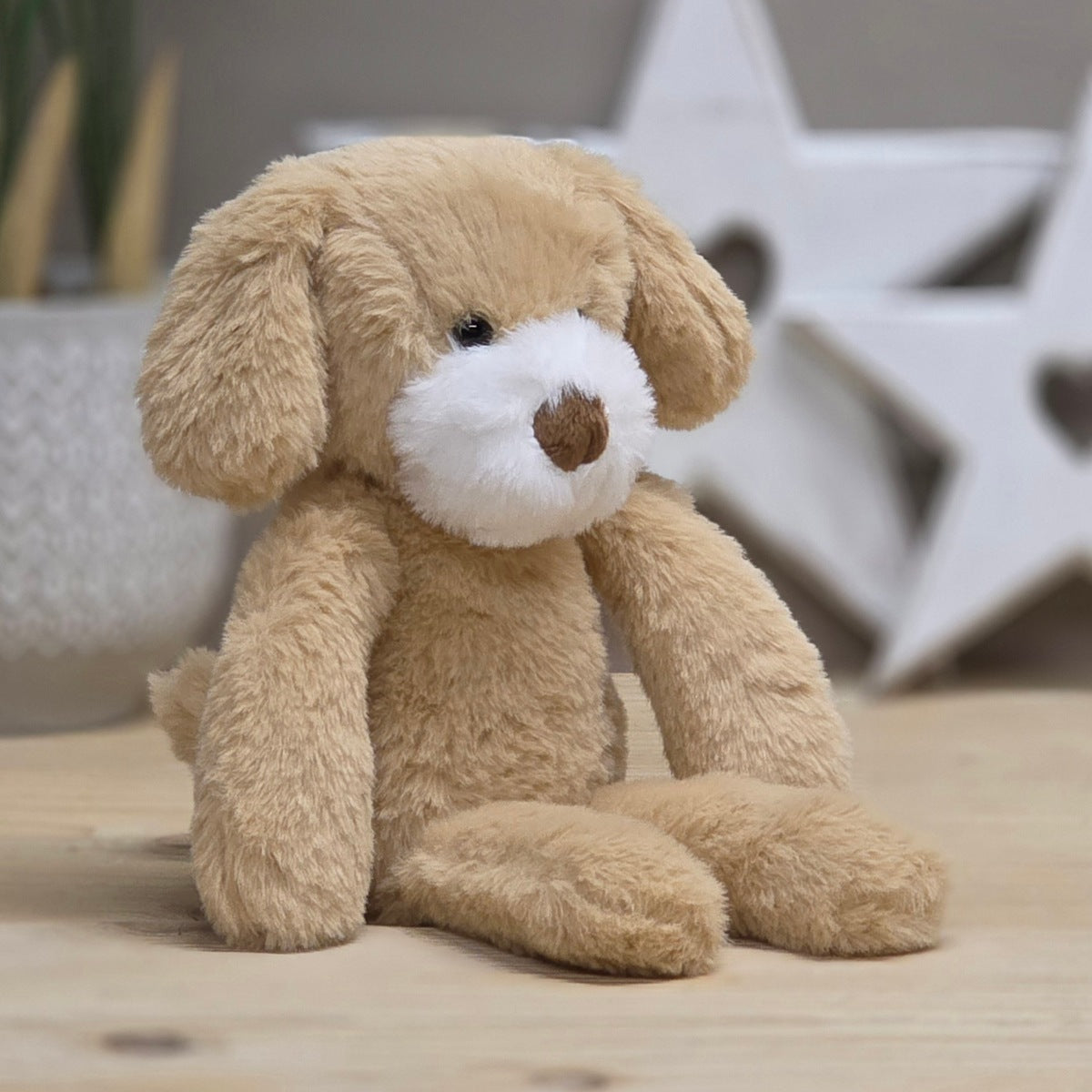 A plush soft stuffed dog toy in light tan with a white snout and brown nose, sitting on a light wood surface with a softly blurred indoor background.