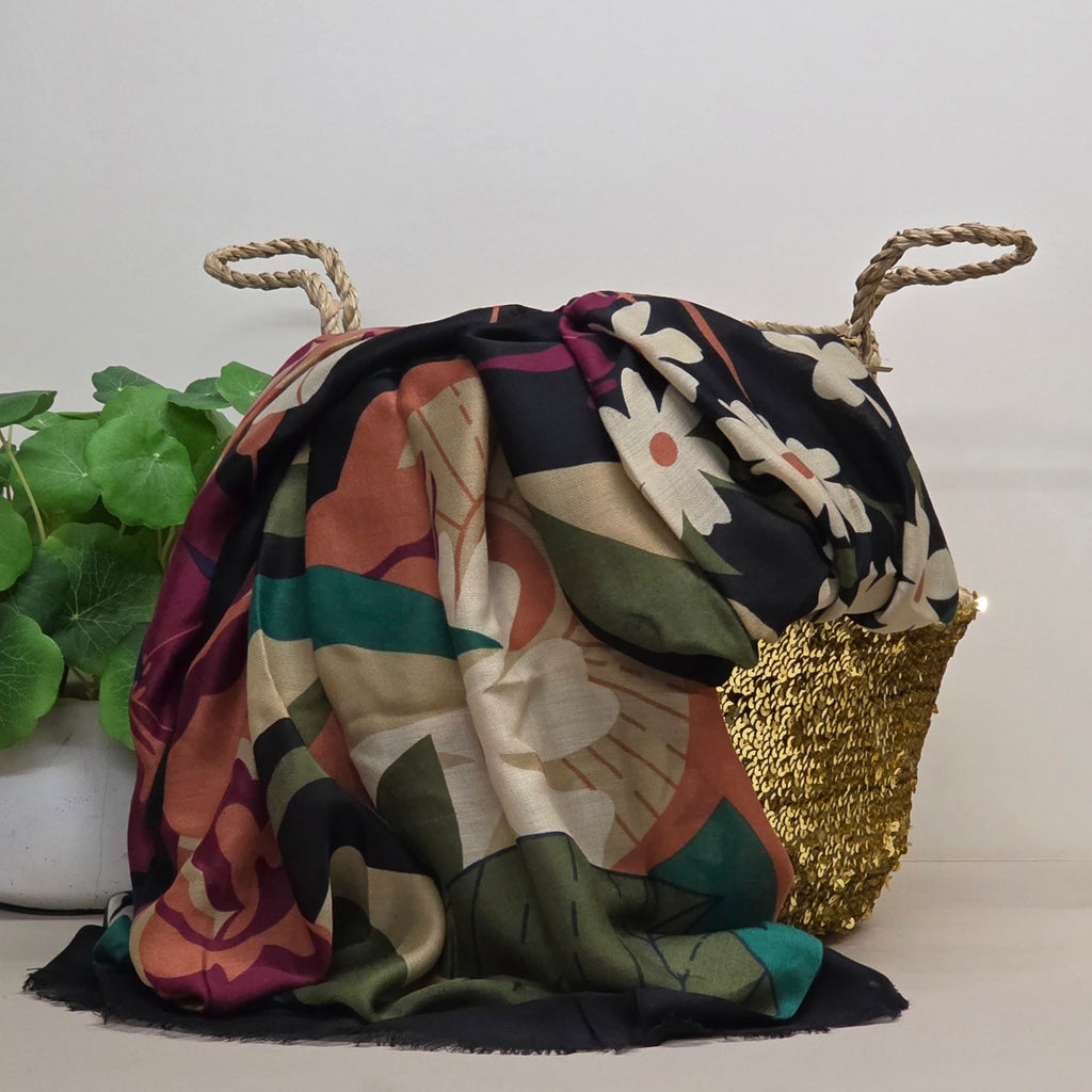 A multicolor floral and abstract patterned scarf draped over a woven straw tote bag with rope handles, set against a plain light background with a green plant on the left.