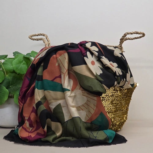 A multicolor floral and abstract patterned scarf draped over a woven straw tote bag with rope handles, set against a plain light background with a green plant on the left.