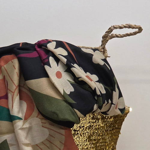 A close-up of a patterned fabric accessory draped over a woven gold basket with a rope handle, against a plain light background.
