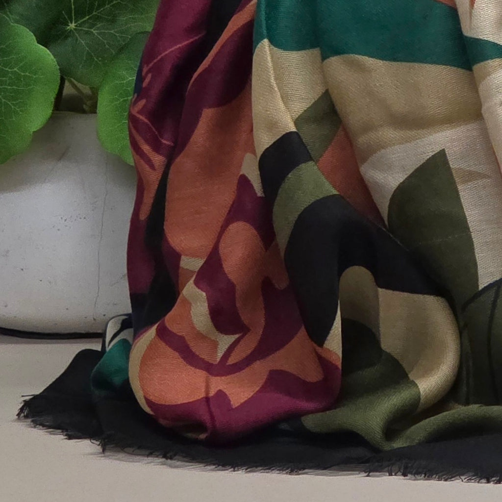 A draped multicolor scarf with a large floral and geometric pattern in burgundy, orange, green, black, and beige, styled next to a white planter with green leaves.