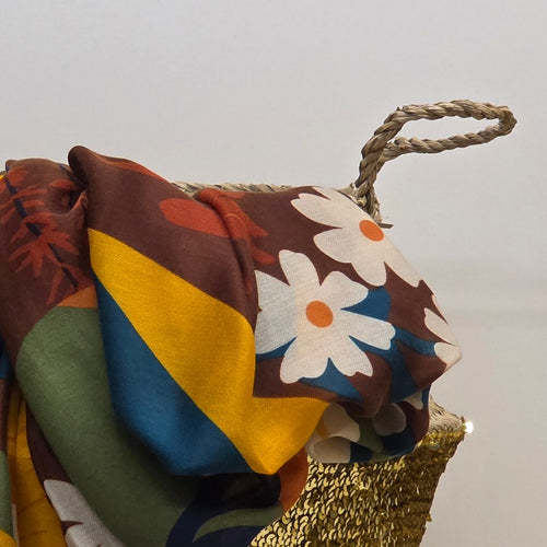 A multicolor patterned scarf draped over a woven straw bag with a rope handle, set against a plain light gray background.