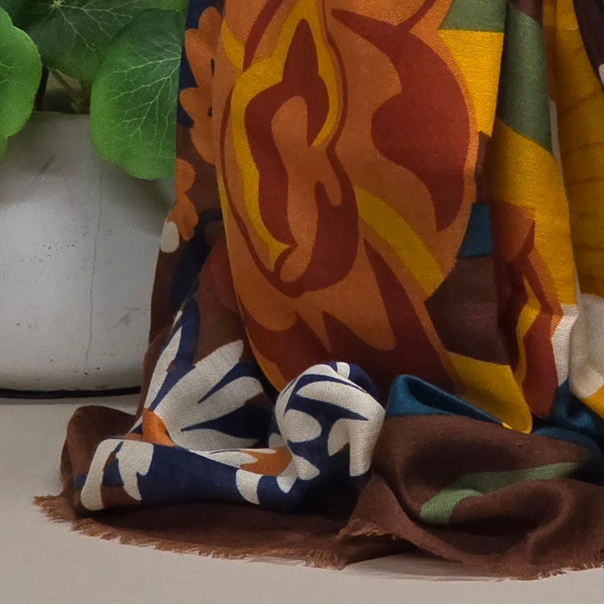 A multicolor patterned scarf draped over a surface, featuring large abstract floral or sunburst shapes in orange, brown, yellow, blue, white, and green. A white planter with green leaves is partially visible in the background.