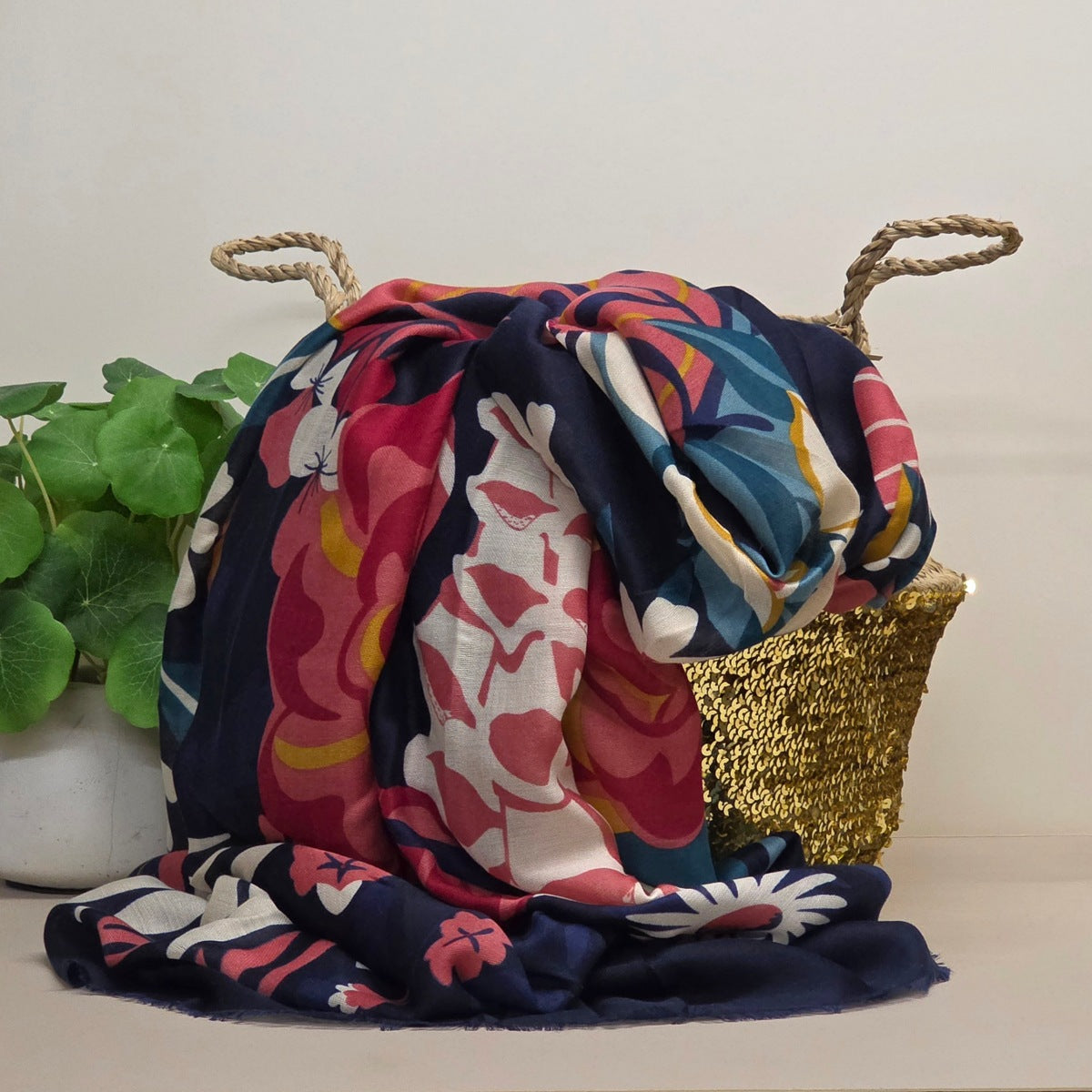 A multicolor floral print scarf draped over a gold woven basket with rope handles, next to a potted green plant, against a plain beige wall.