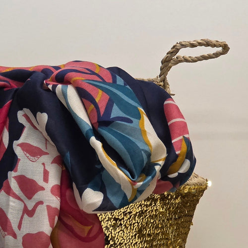 A multicolor tropical print scarf draped over a woven gold chair, featuring navy blue, pink, teal, yellow, and white patterns.