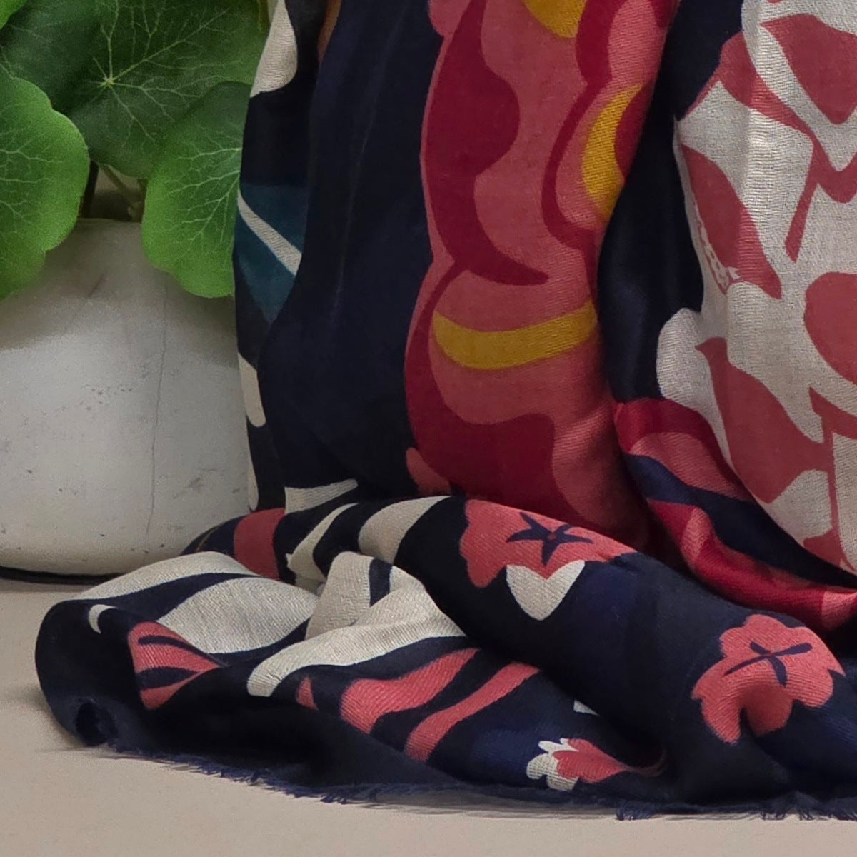 A folded patterned scarf draped on a surface next to a white planter with green leaves. The scarf has a navy blue base with abstract floral shapes in pink, red, yellow, and off-white.
