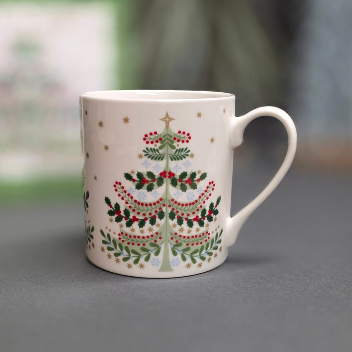 A white ceramic mug featuring a hand-painted Christmas tree design with green holly leaves, red berries, and gold stars, set against a blurred grey and green background.