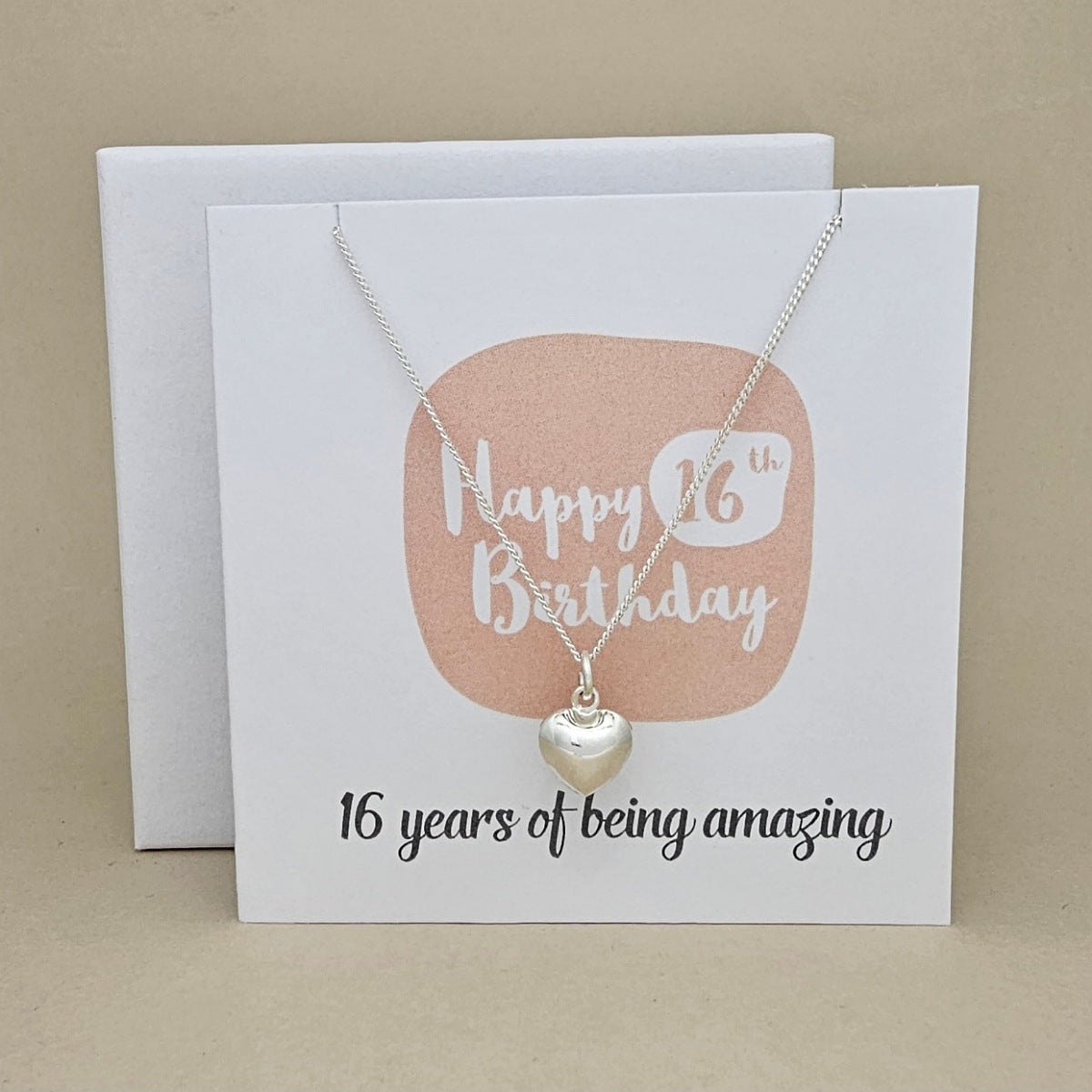A silver heart necklace displayed on a white card with a peach rounded square and the text 'Happy 16th Birthday' and ’16 years of being amazing,' 
