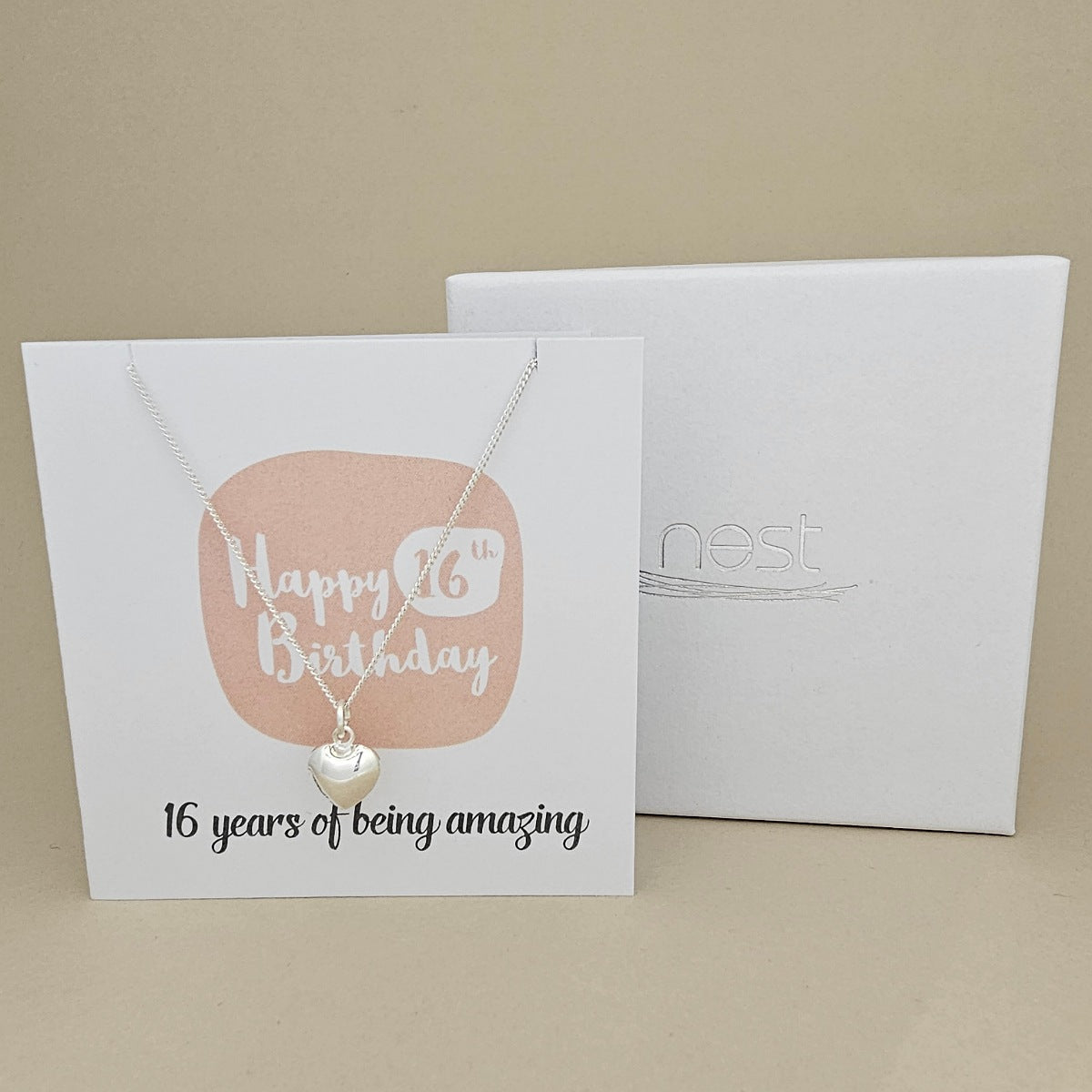 A silver heart necklace displayed on a white card with a peach rounded square and the text 'Happy 16th Birthday' and ’16 years of being amazing,' shown in front of a white branded 'nest' gift box on a beige background.
