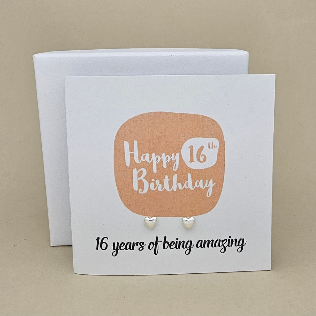 A pair of silver heart earrings displayed on a white card with a peach rounded square and the text 'Happy 16th Birthday' and ’16 years of being amazing,' 
