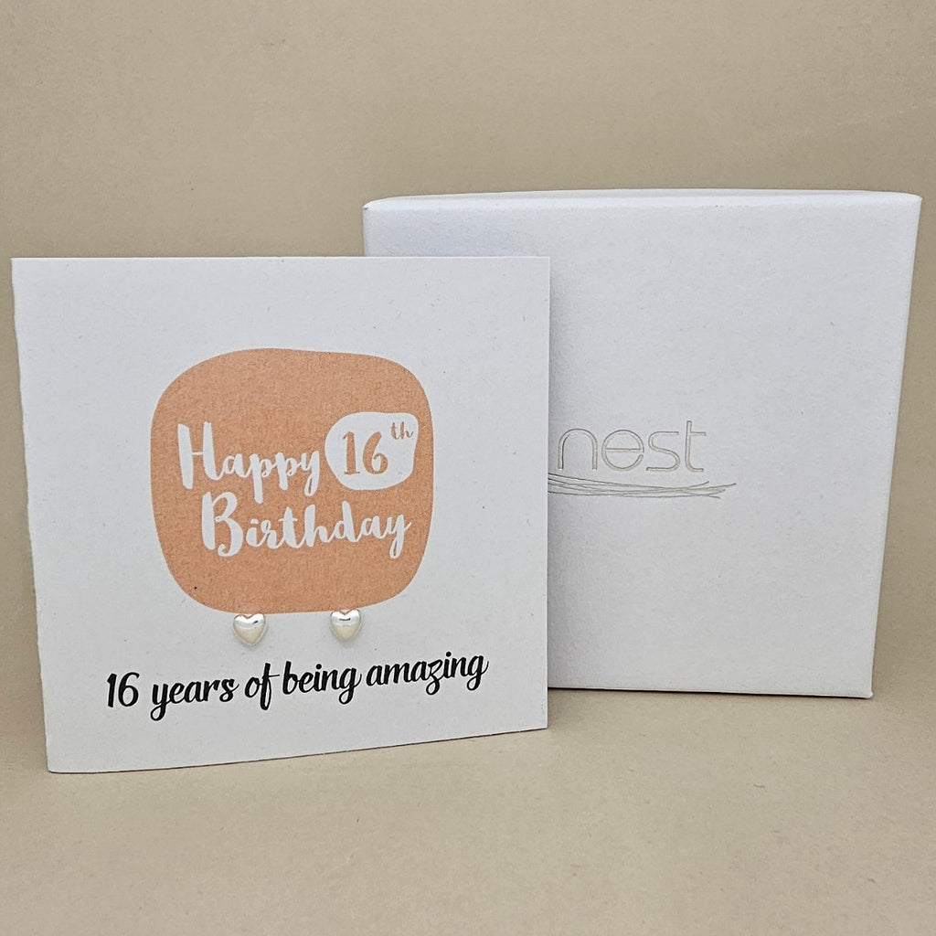 A pair of silver heart earrings displayed on a white card with a peach rounded square and the text 'Happy 16th Birthday' and ’16 years of being amazing,' shown in front of a white branded 'nest' gift box on a beige background.
