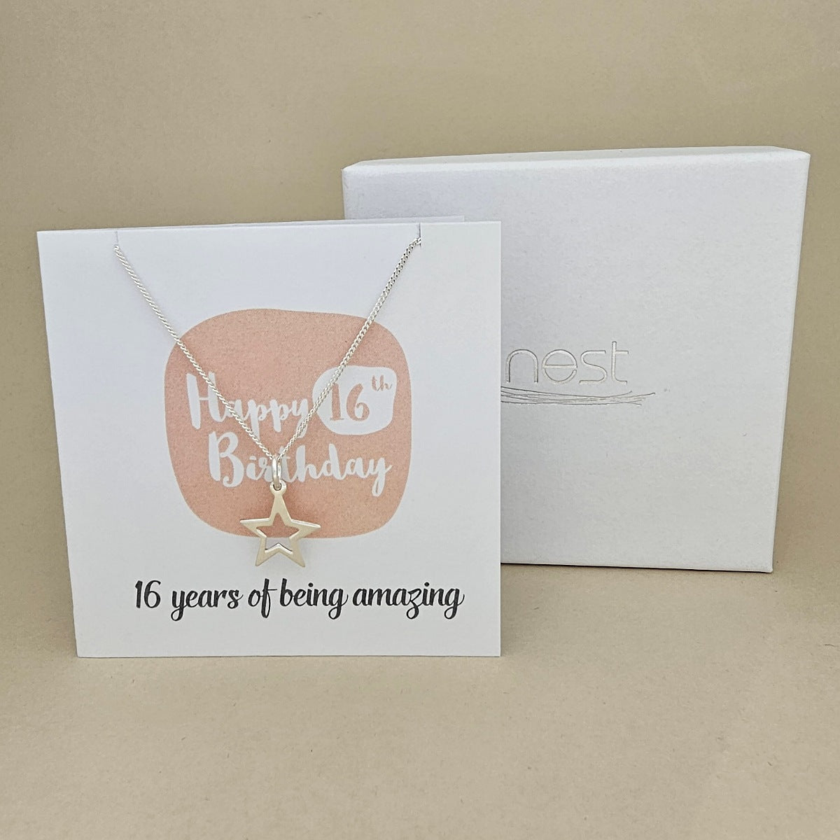 A silver open star necklace displayed on a white card with a peach rounded square and the text 'Happy 16th Birthday' and ’16 years of being amazing,' shown in front of a white branded 'nest' gift box on a beige background.
