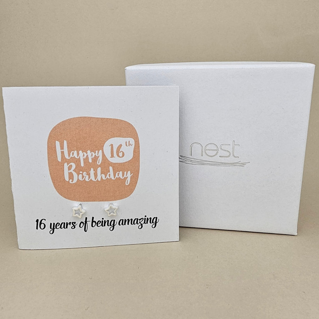 A pair of silver open star earrings displayed on a white card with a peach rounded square and the text 'Happy 16th Birthday' and ’16 years of being amazing,' shown in front of a white branded 'nest' gift box on a beige background
