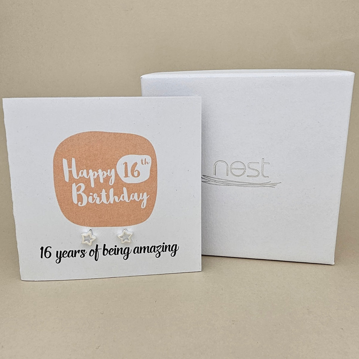 A pair of silver open star earrings displayed on a white card with a peach rounded square and the text 'Happy 16th Birthday' and ’16 years of being amazing,' shown in front of a white branded 'nest' gift box on a beige background
