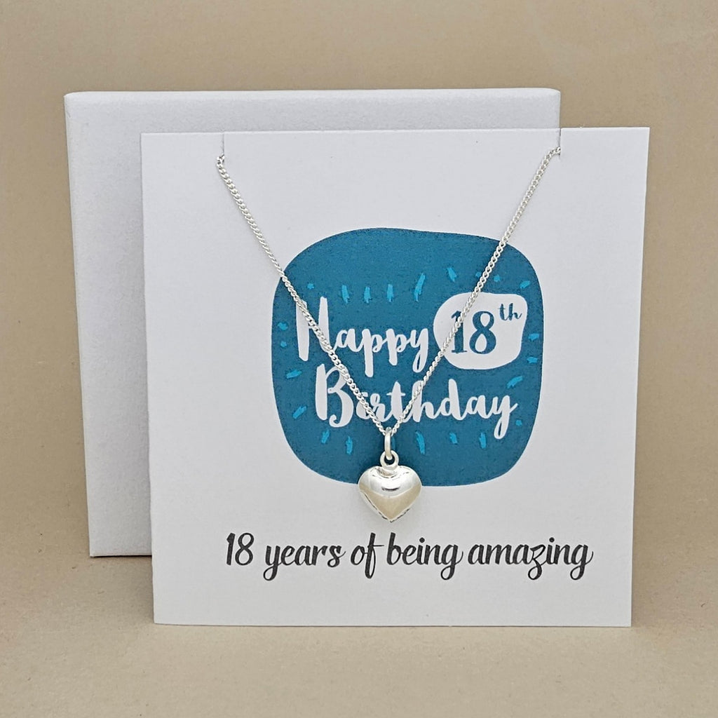 A silver heart necklace displayed on a white card with a blue rounded square and the text 'Happy 18th Birthday' and ’18 years of being amazing,'
