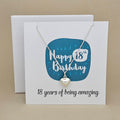 A silver heart necklace displayed on a white card with a blue rounded square and the text 'Happy 18th Birthday' and ’18 years of being amazing,'
