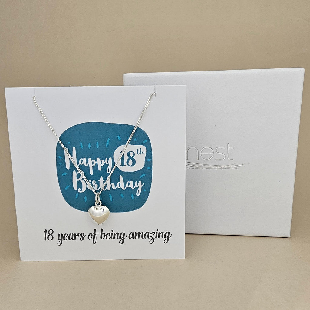 A silver heart necklace displayed on a white card with a blue rounded square and the text 'Happy 18th Birthday' and ’18 years of being amazing,' shown in front of a white branded 'nest' gift box on a beige background.
