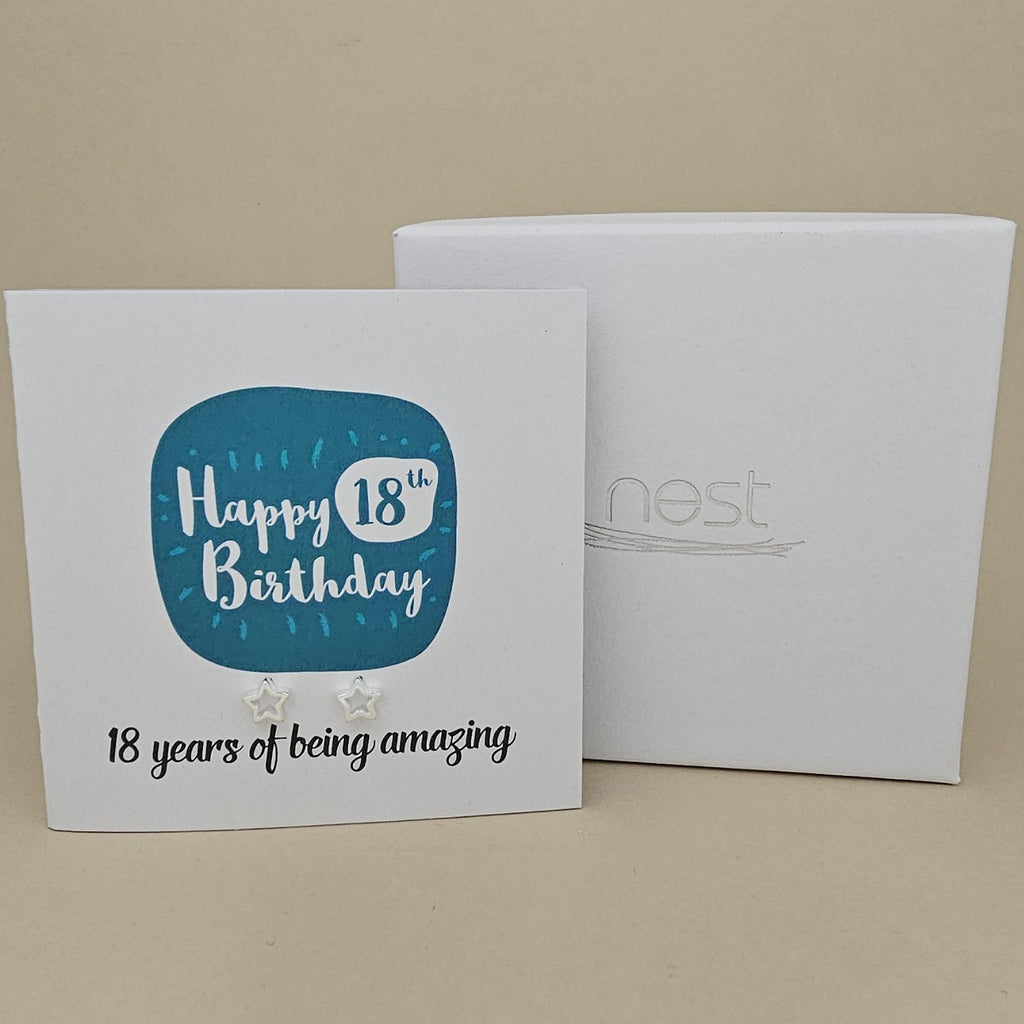 A pair of silver open star earrings displayed on a white card with a blue rounded square and the text 'Happy 18th Birthday' and ’18 years of being amazing,' shown in front of a white branded 'nest' gift box on a beige background
