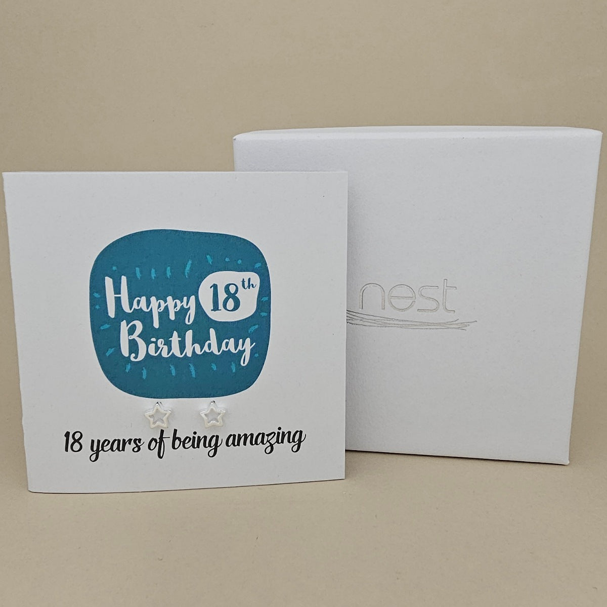 A pair of silver open star earrings displayed on a white card with a blue rounded square and the text 'Happy 18th Birthday' and ’18 years of being amazing,' shown in front of a white branded 'nest' gift box on a beige background
