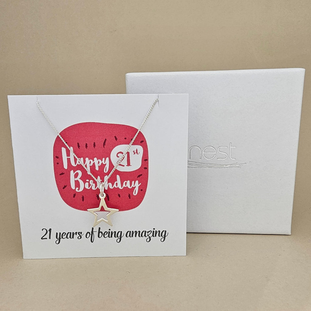A silver open star necklace displayed on a white card with a bright pink rounded square and the text 'Happy 21st Birthday' and ’21 years of being amazing,' shown in front of a white branded 'nest' gift box on a beige background.
