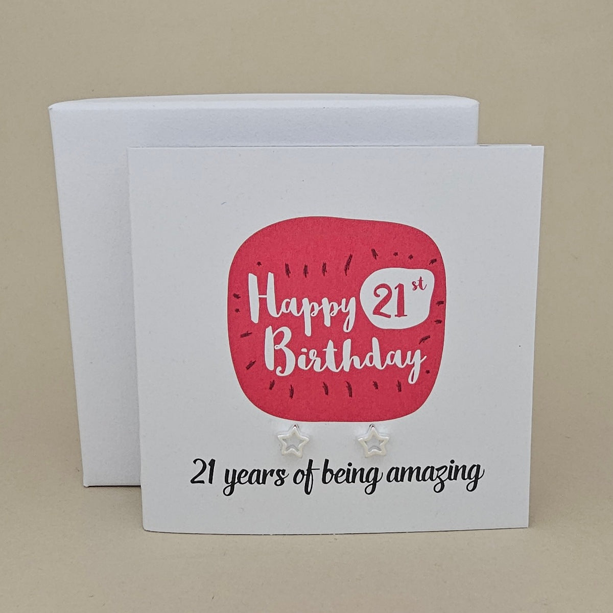 A pair of silver open star earrings displayed on a white card with a bright pink rounded square and the text 'Happy 21st Birthday' and ’21 years of being amazing,' 
