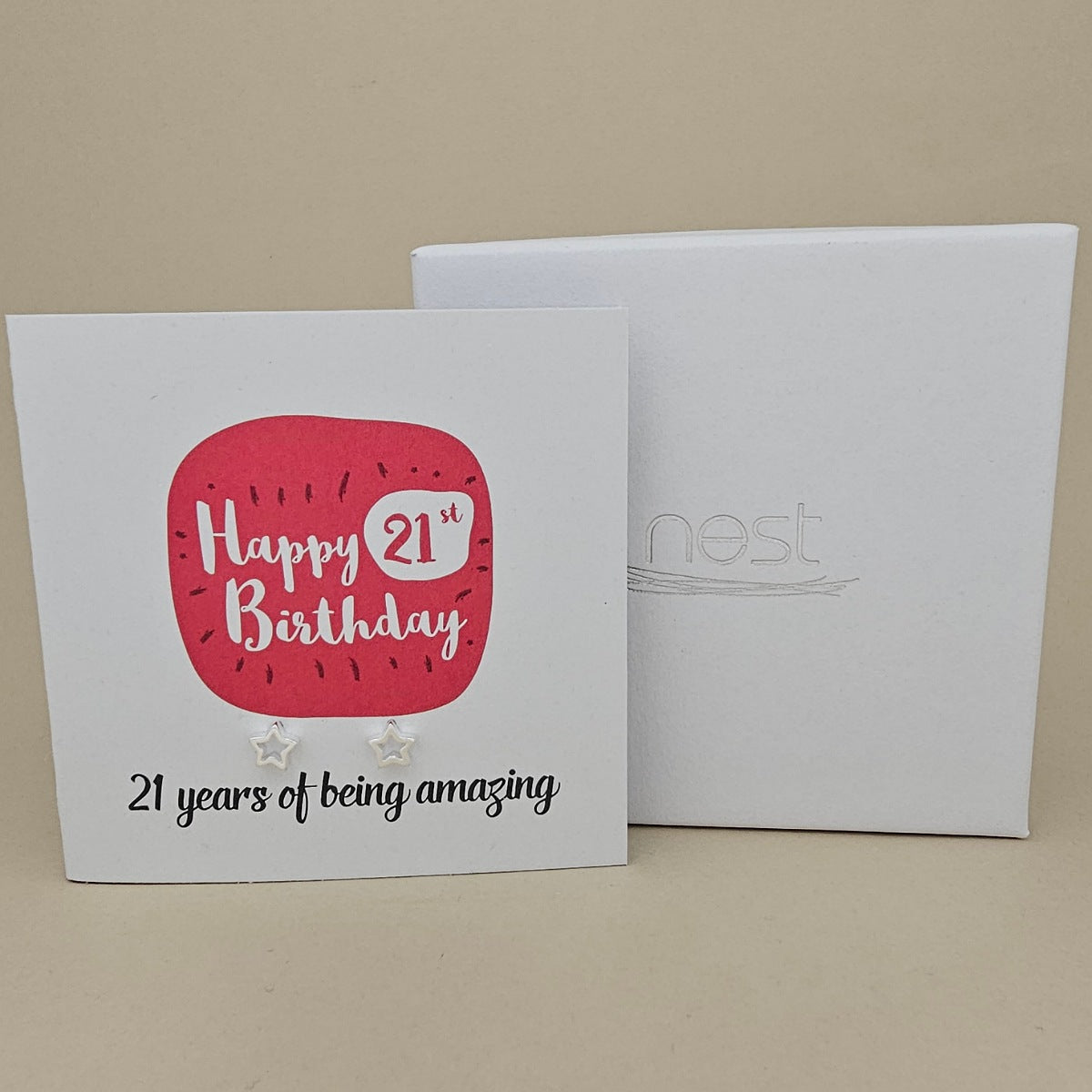 A pair of silver open star earrings displayed on a white card with a bright pink rounded square and the text 'Happy 21st Birthday' and ’21 years of being amazing,' shown in front of a white branded 'nest' gift box on a beige background
