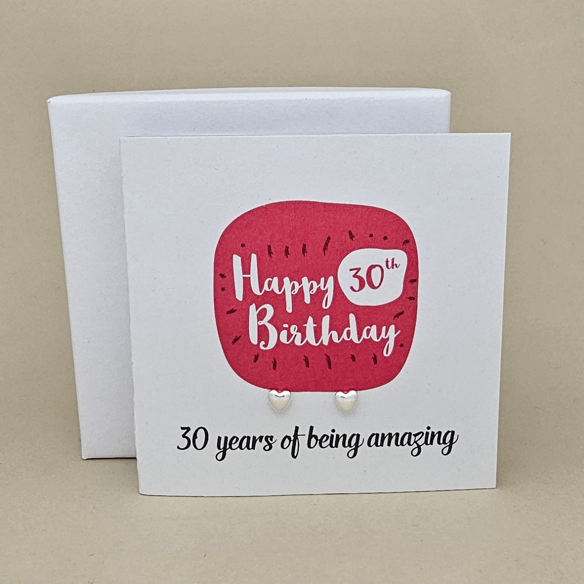 A pair of silver heart earrings displayed on a white card with a bright pink rounded square and the text 'Happy 30th Birthday' and ’30 years of being amazing,' 

