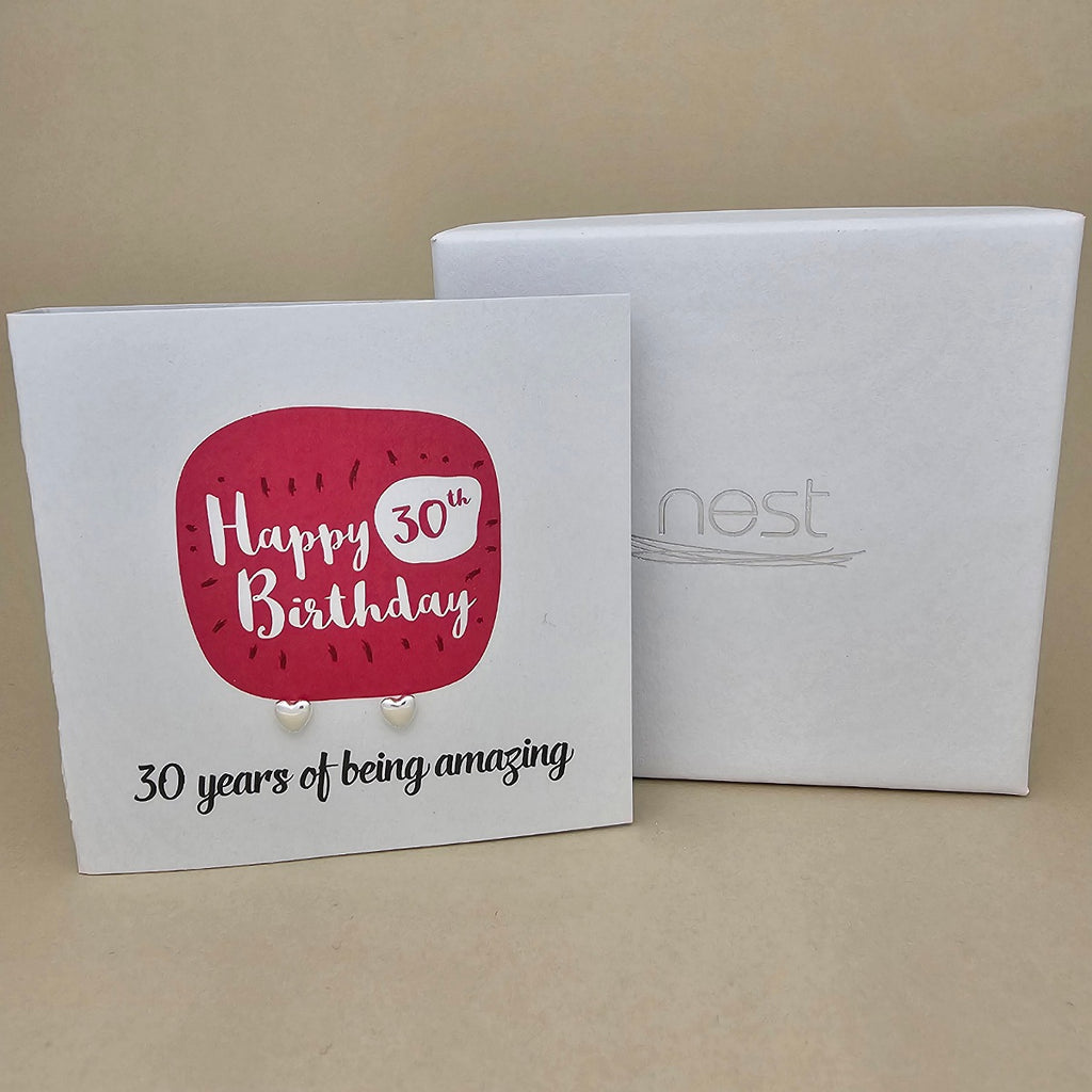 A pair of silver heart earrings displayed on a white card with a bright pink rounded square and the text 'Happy 30th Birthday' and ’30 years of being amazing,' shown in front of a white branded 'nest' gift box on a beige background.
