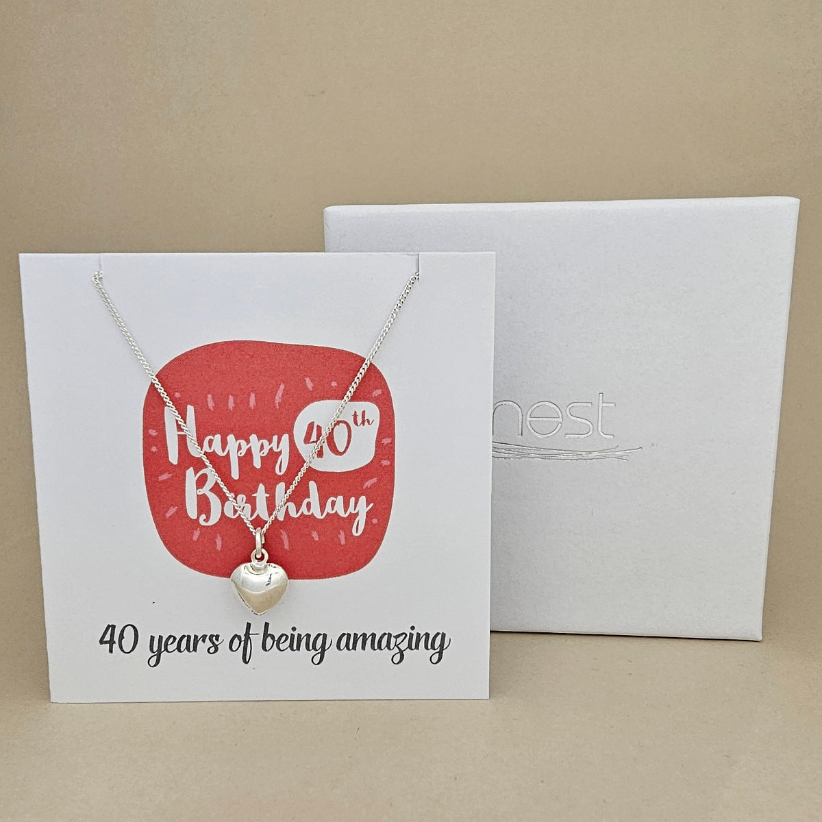 A silver heart necklace displayed on a white card with a red rounded square and the text 'Happy 40th Birthday' and ’40 years of being amazing,' shown in front of a white branded 'nest' gift box on a beige background.
