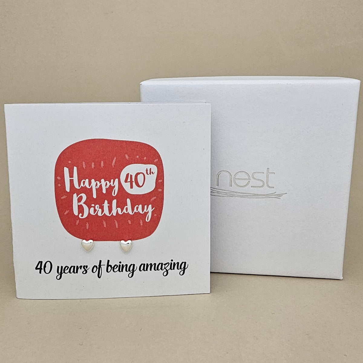 A pair of silver heart earrings displayed on a white card with a red rounded square and the text 'Happy 40th Birthday' and ’40 years of being amazing,' shown in front of a white branded 'nest' gift box on a beige background.
