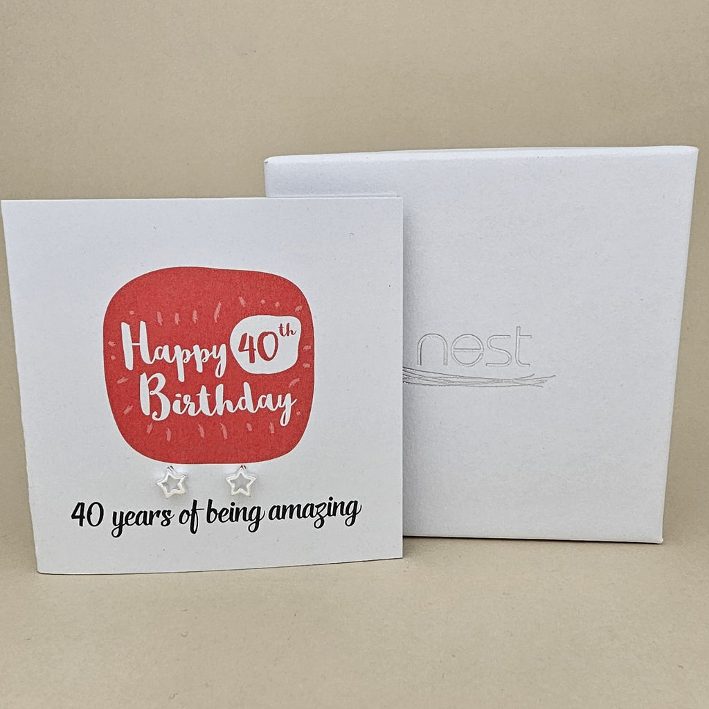 A pair of silver open star earrings displayed on a white card with a red rounded square and the text 'Happy 40th Birthday' and ’40 years of being amazing,' shown in front of a white branded 'nest' gift box on a beige background
