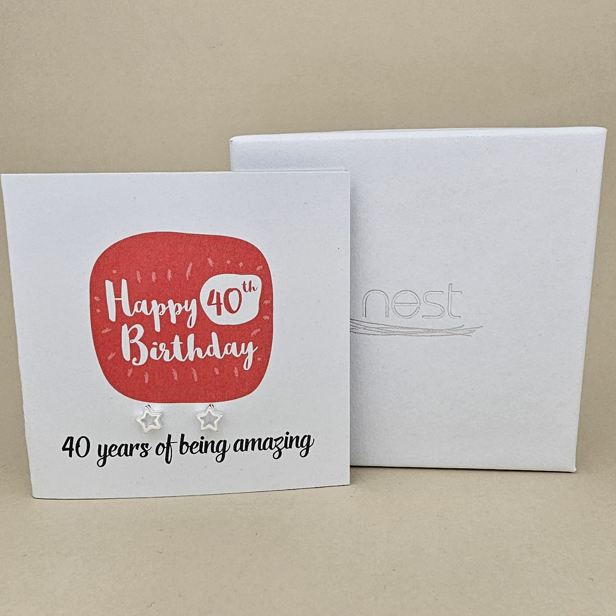 A pair of silver open star earrings displayed on a white card with a red rounded square and the text 'Happy 40th Birthday' and ’40 years of being amazing,' shown in front of a white branded 'nest' gift box on a beige background
