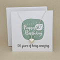 A silver heart necklace displayed on a white card with a green rounded square and the text 'Happy 50th Birthday' and ’50 years of being amazing,' 
