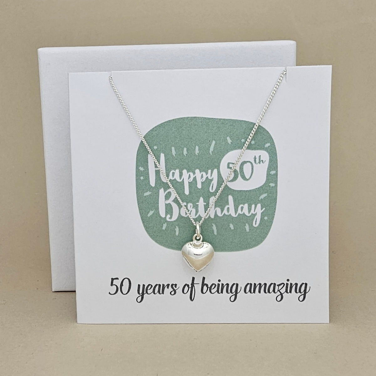 A silver heart necklace displayed on a white card with a green rounded square and the text 'Happy 50th Birthday' and ’50 years of being amazing,' 
