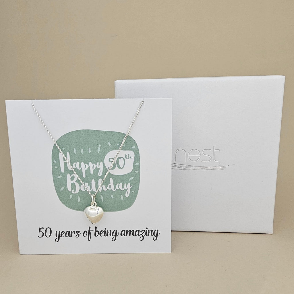 A silver heart necklace displayed on a white card with a green rounded square and the text 'Happy 50th Birthday' and ’50 years of being amazing,' shown in front of a white branded 'nest' gift box on a beige background.
