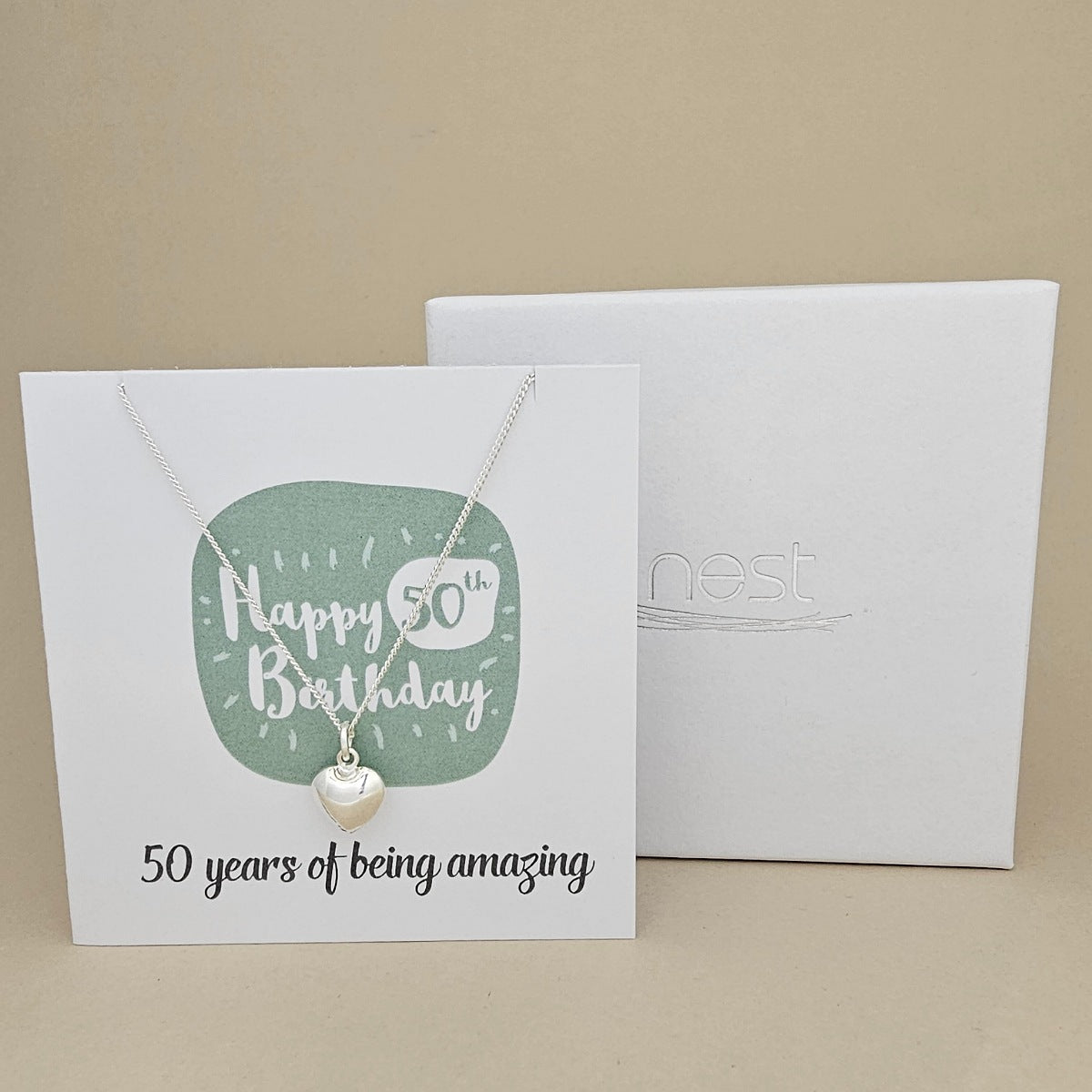 A silver heart necklace displayed on a white card with a green rounded square and the text 'Happy 50th Birthday' and ’50 years of being amazing,' shown in front of a white branded 'nest' gift box on a beige background.
