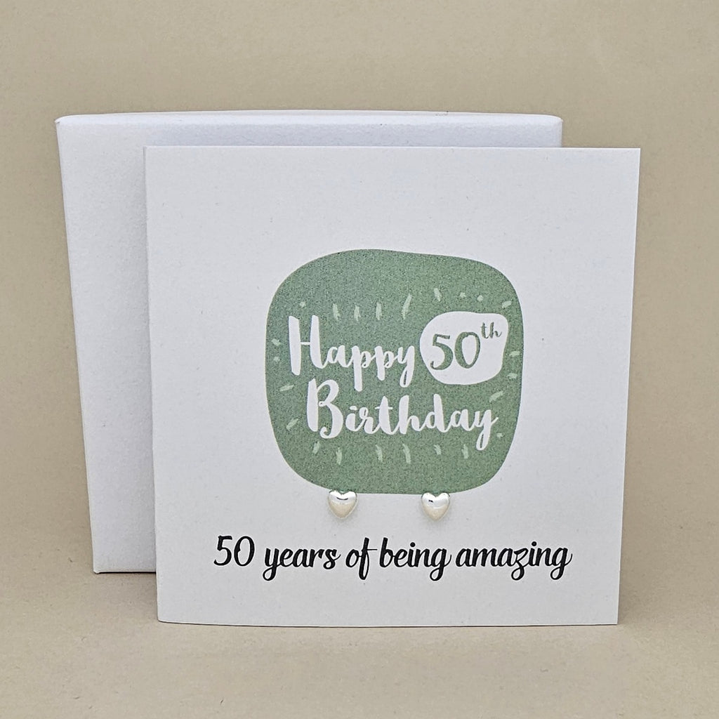 A pair of silver heart earrings displayed on a white card with a green rounded square and the text 'Happy 50th Birthday' and ’50 years of being amazing,' 
