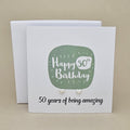 A pair of silver heart earrings displayed on a white card with a green rounded square and the text 'Happy 50th Birthday' and ’50 years of being amazing,' 
