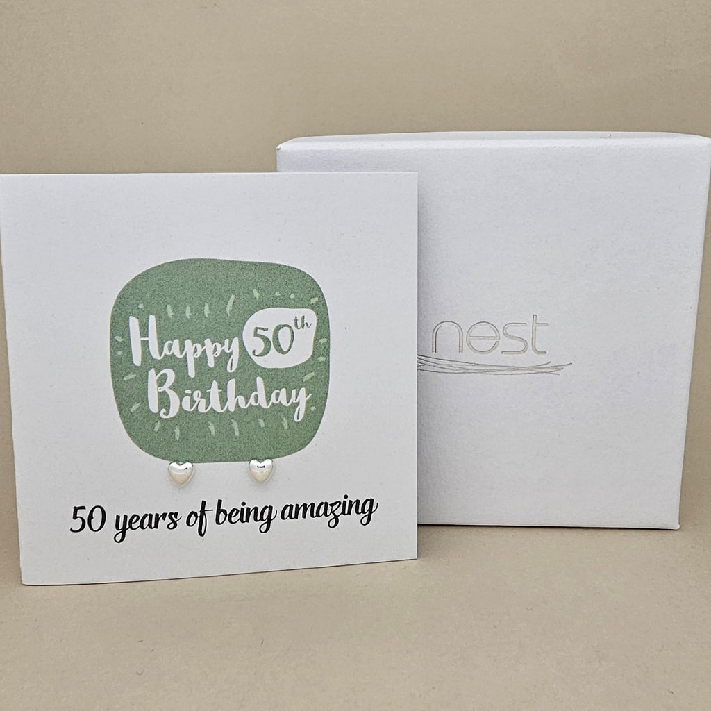 A pair of silver heart earrings displayed on a white card with a green rounded square and the text 'Happy 50th Birthday' and ’50 years of being amazing,' shown in front of a white branded 'nest' gift box on a beige background.
