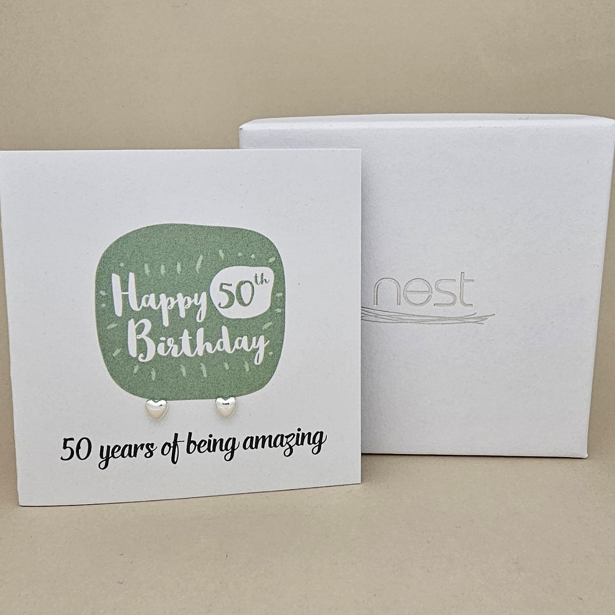 A pair of silver heart earrings displayed on a white card with a green rounded square and the text 'Happy 50th Birthday' and ’50 years of being amazing,' shown in front of a white branded 'nest' gift box on a beige background.
