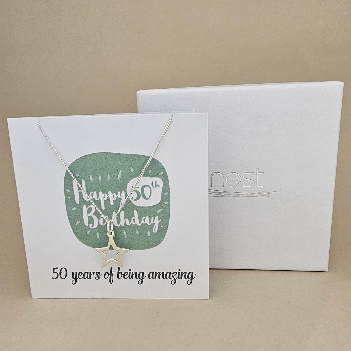 A silver open star necklace displayed on a white card with a green rounded square and the text 'Happy 50th Birthday' and ’50 years of being amazing,' shown in front of a white branded 'nest' gift box on a beige background.
