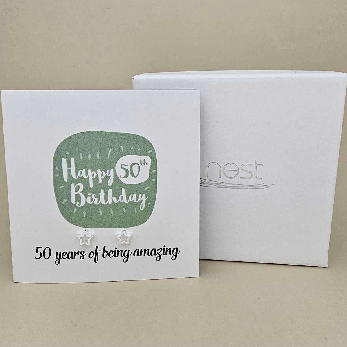 A pair of silver open star earrings displayed on a white card with a green rounded square and the text 'Happy 50th Birthday' and ’50 years of being amazing,' shown in front of a white branded 'nest' gift box on a beige background
