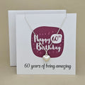 A silver heart necklace displayed on a white card with a burgundy rounded square and the text 'Happy 60th Birthday' and '60 years of being amazing,'.
