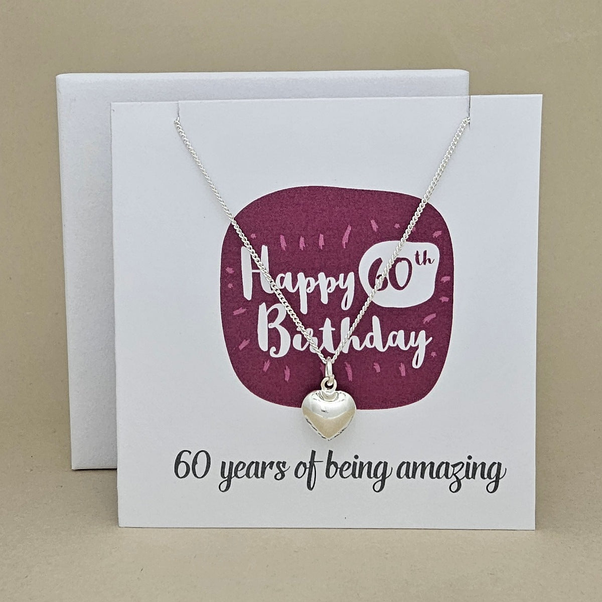 A silver heart necklace displayed on a white card with a burgundy rounded square and the text 'Happy 60th Birthday' and '60 years of being amazing,'.
