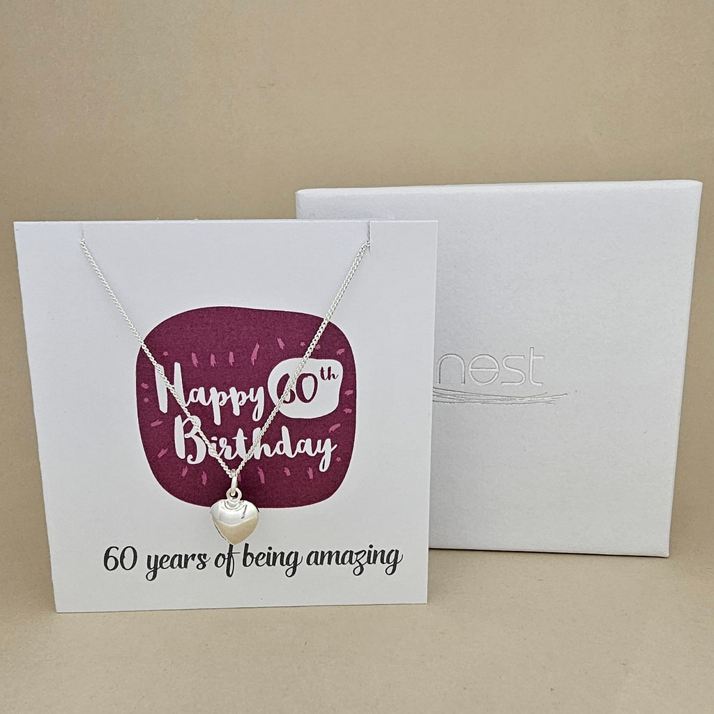 A silver heart necklace displayed on a white card with a burgundy rounded square and the text 'Happy 60th Birthday' and '60 years of being amazing,' shown in front of a white branded 'nest' gift box on a beige background.