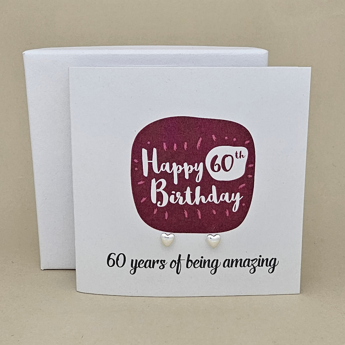 A pair of silver heart earrings displayed on a white card with a burgundy rounded square and the text 'Happy 60th Birthday' and '60 years of being amazing,'.