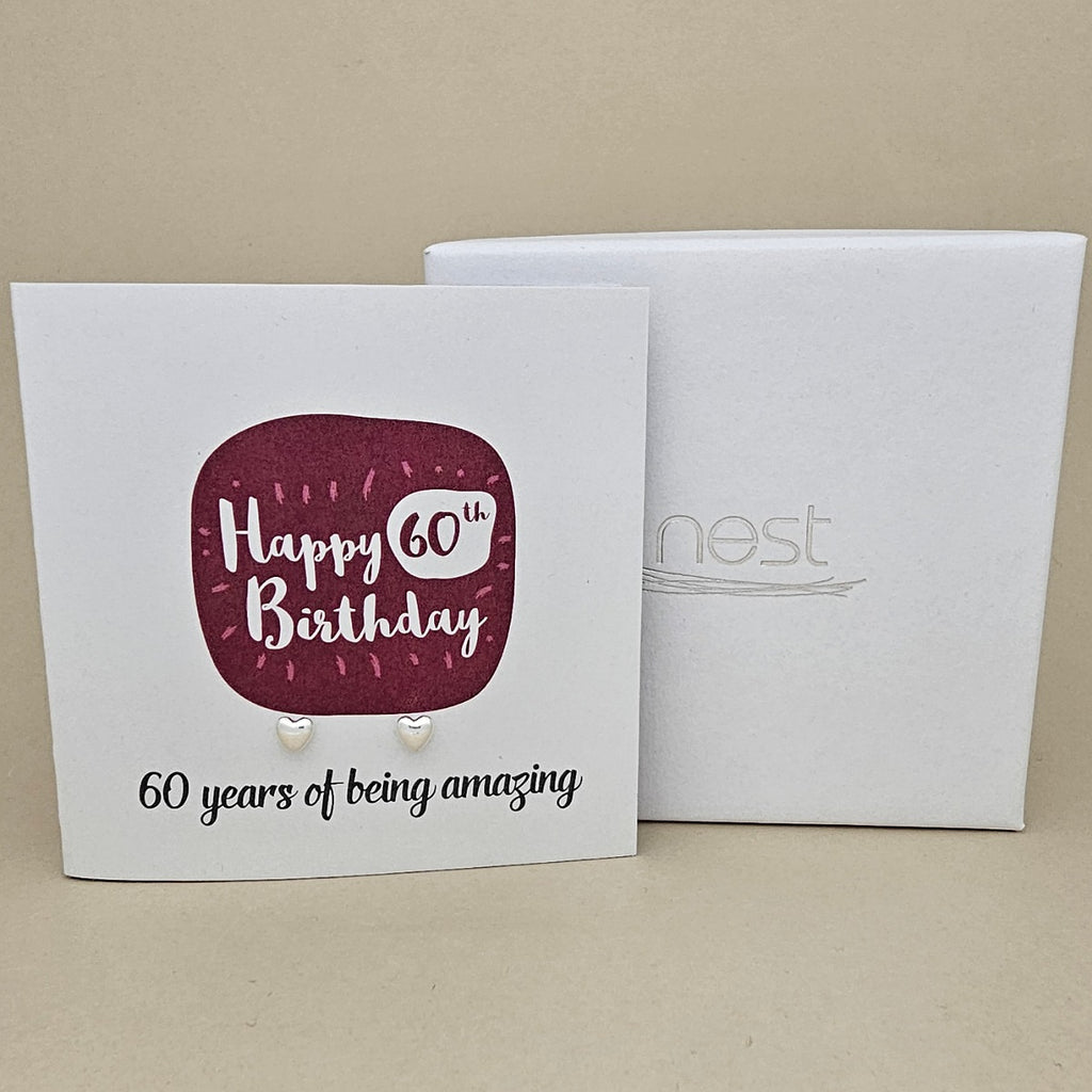 A pair of silver heart earrings displayed on a white card with a burgundy rounded square and the text 'Happy 60th Birthday' and '60 years of being amazing,' shown in front of a white branded 'nest' gift box on a beige background.